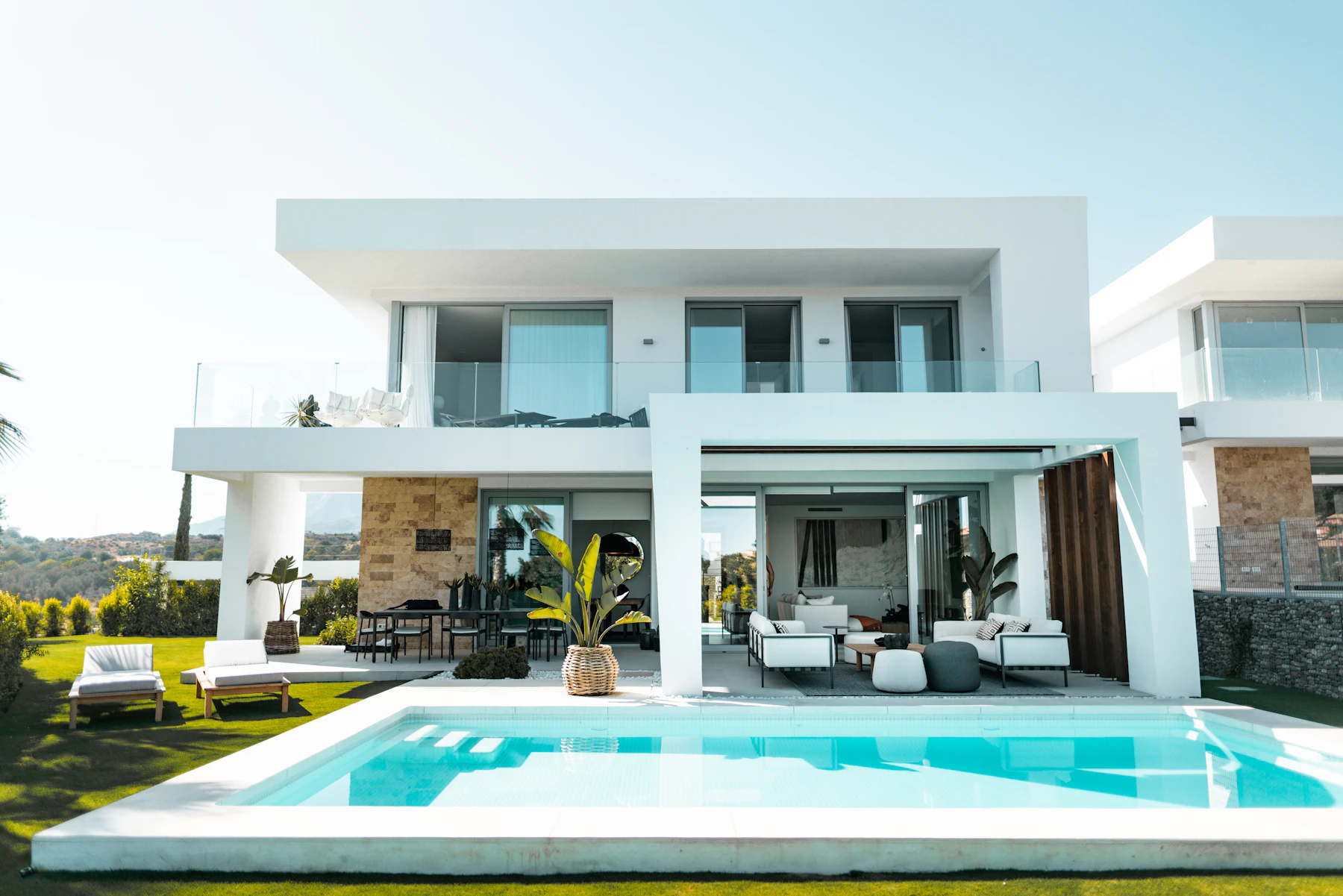 Completed luxury villa at dusk: white cantilevered upper storey over full-height
glazing, stone-clad walls, an infinity pool and planted terrace